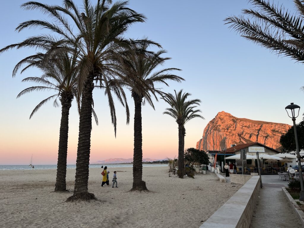 Palmen am Strand von San Vito Lo Capo vor dem orange beleuchteten Monte Monaco bei Sonnenuntergang an der sizilianischen Küste.