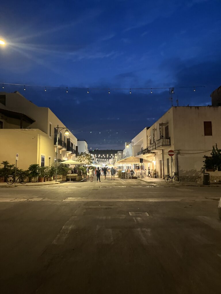 Belebte Fußgängerzone in San Vito Lo Capo am Abend mit Lichterketten, Restaurants, historischen Gebäuden und Menschen beim Flanieren.