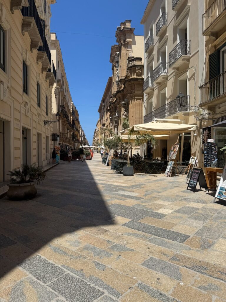 Corso Vittorio Emanuele in Trapani mit historischen Gebäuden, Cafés, Restaurants und gepflasterter Fußgängerzone unter blauem Himmel.