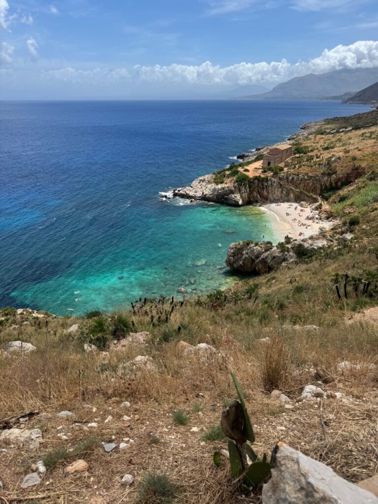 Blick von einem Hang auf die Cala Tonnarella dell’Uzzo im Naturreservat Zingaro mit türkisfarbenem Wasser, kleiner Kiesbucht und felsiger Küstenlandschaft.