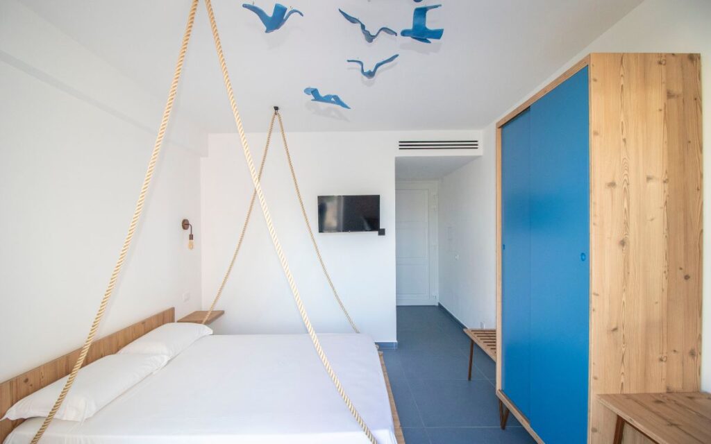Modernes Hotelzimmer im Hotel ARTEMARe in San Vito Lo Capo mit großem Bett, blauen Designelementen, Holzmöbeln und maritimer Dekoration an der Decke.