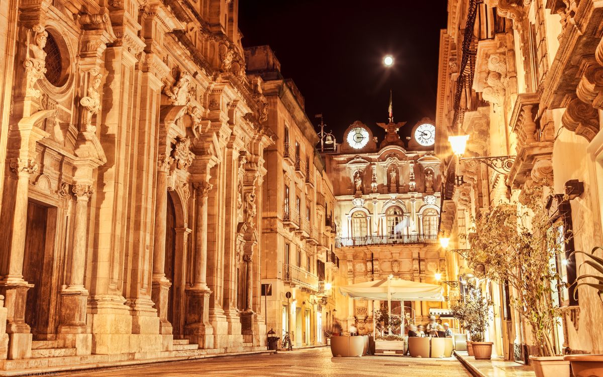 Beleuchteter Palazzo Senatorio mit Torre dell’Orologio in der Altstadt von Trapani bei Nacht, umgeben von barocken Gebäuden und einer historischen Gasse.