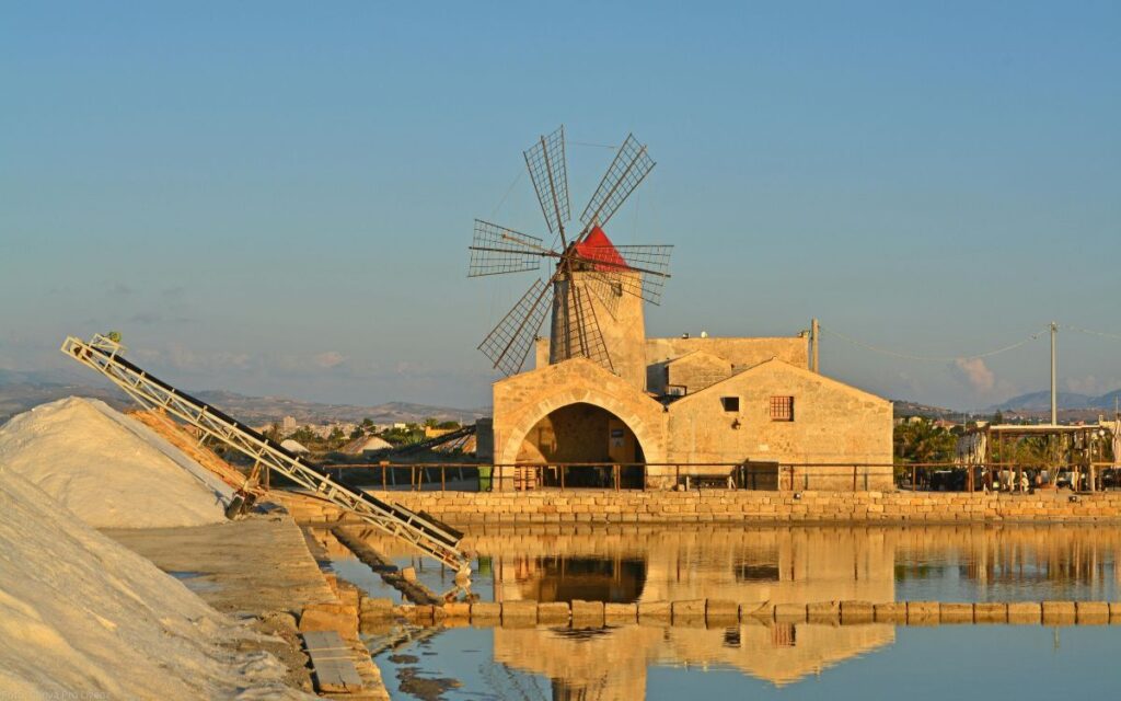 Historische Windmühle der Salinen von Trapani mit Salzbecken und reflektierendem Wasser unter klarem Himmel in Westsizilien.