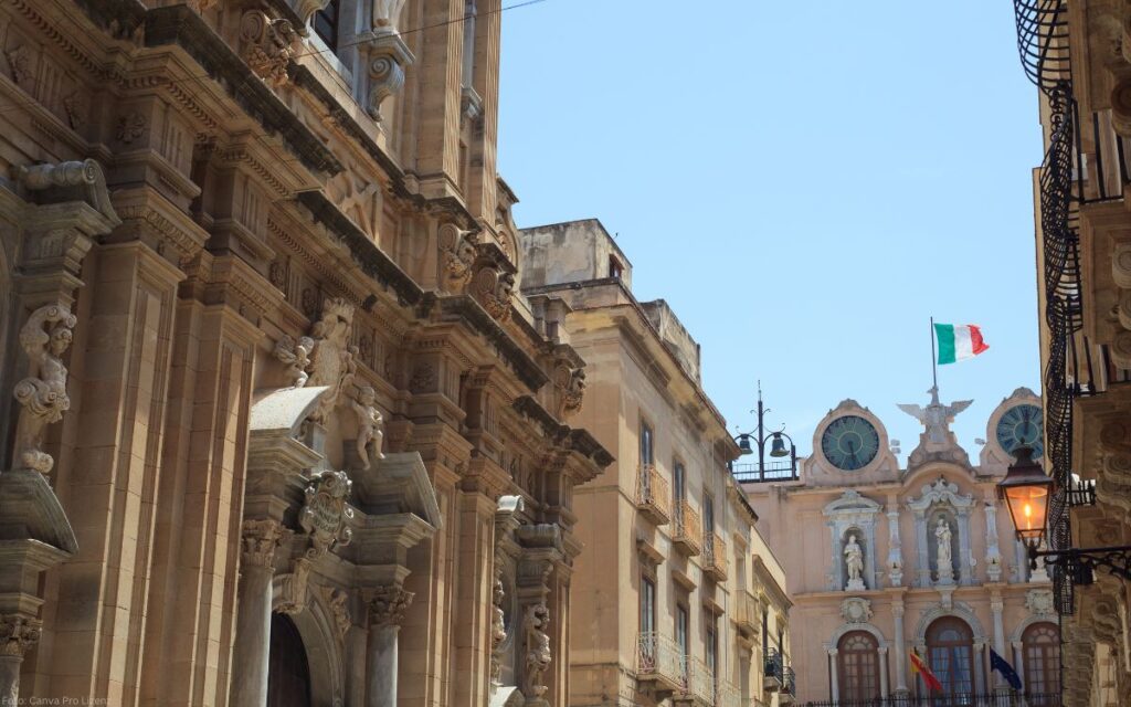 Historische Straße in der Altstadt von Trapani mit barocken Gebäuden, Kirchenfassaden und italienischer Flagge unter blauem Himmel.